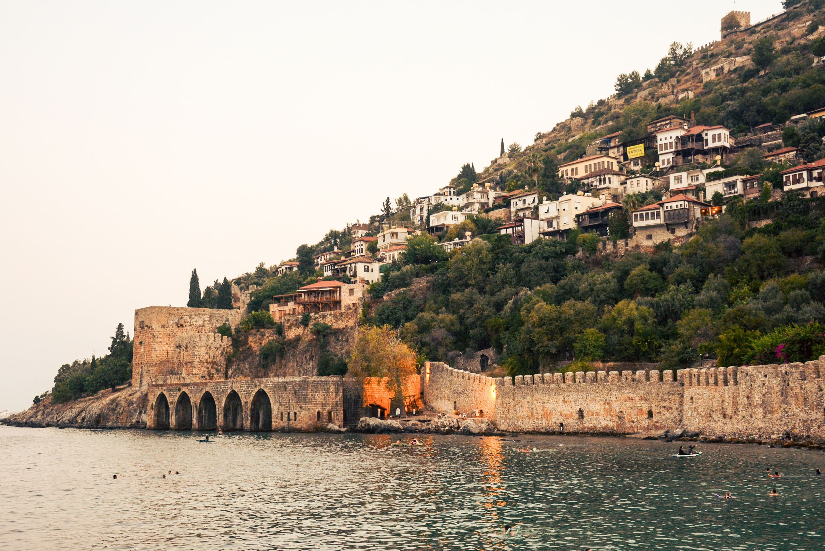 Alanya Tersanesi'nin kemerli taş kapıları ve kale surları denize bakıyor, arkasında yamaçtaki beyaz evler yükseliyor, denizde yüzen misafirler var
