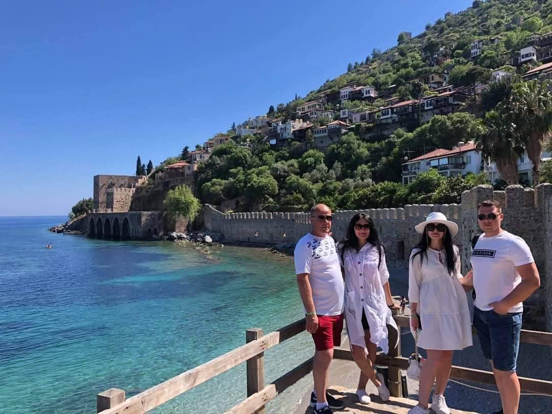 Dört ziyaretçi Alanya Kalesi'nin sur duvarında duruyor, altta turkuaz koy ve kıyıya kurulmuş eski evler