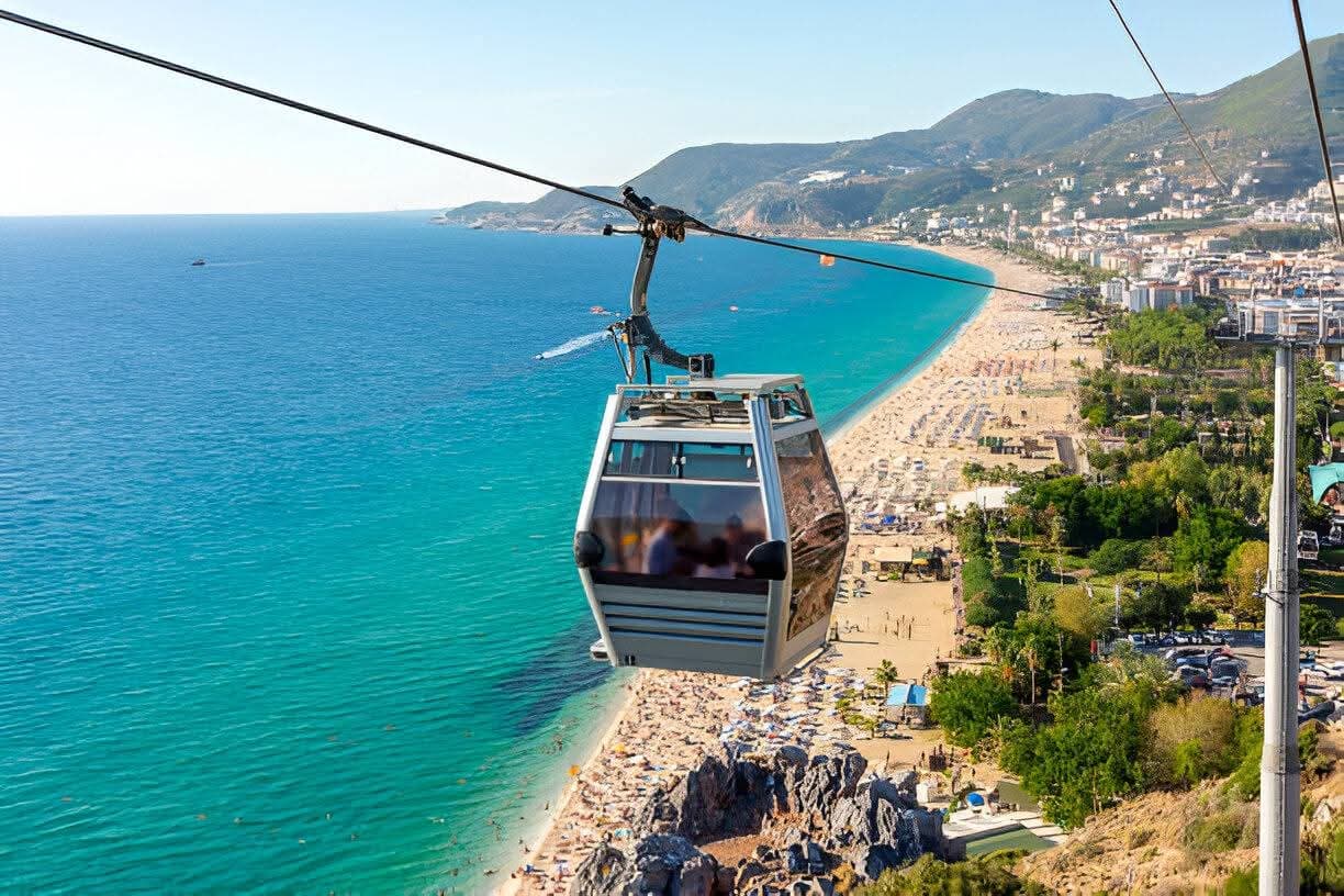 Teleferik kabini Kleopatra Plajı'nın üzerinden geçiyor, altta sahil boyunca dizilmiş şemsiyeler ve turkuaz deniz