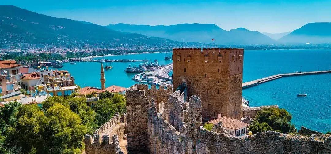 Alanya Kalesi'nin Kızıl Kule'si ve tersanesi kuş bakışı görünüyor, arkada liman, şehir ve dağlar