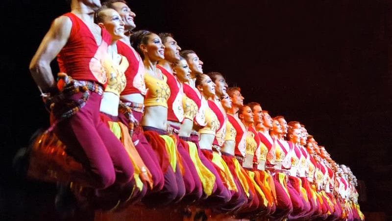 Alev desenli kostümlü onlarca dansçı sahnede tek sıra hâlinde havaya zıplıyor, karanlık sahnede kırmızı ışıklandırma