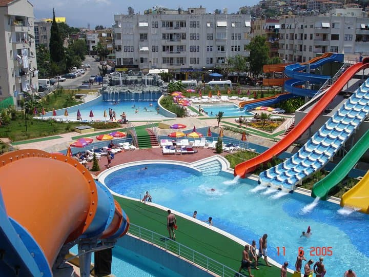 Kuşbakışı görünüm: turuncu, kırmızı, sarı ve yeşil su kaydırakları havuzlara dökülüyor, arkada şehir binaları ve dağlar