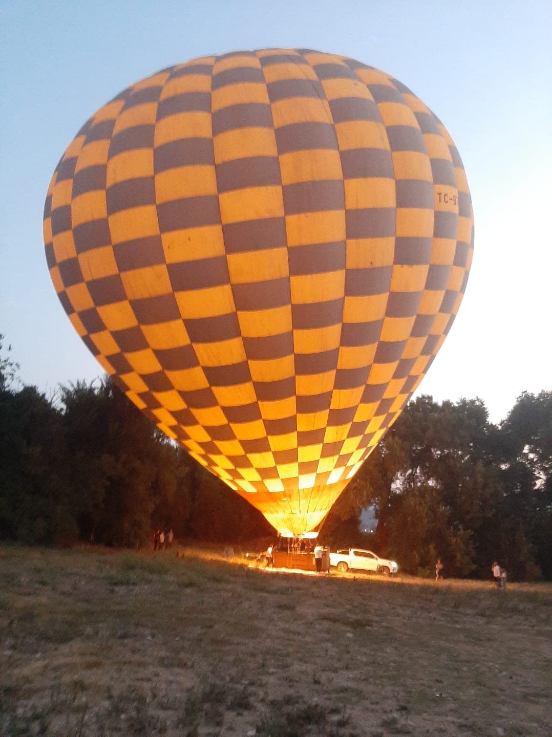 Sarı-gri damalı desenli bir sıcak hava balonu sabahın alacakaranlığında hazırlanıyor, içindeki alevle aydınlanıyor; yanında beyaz bir pikap ve bekleyen birkaç kişi var