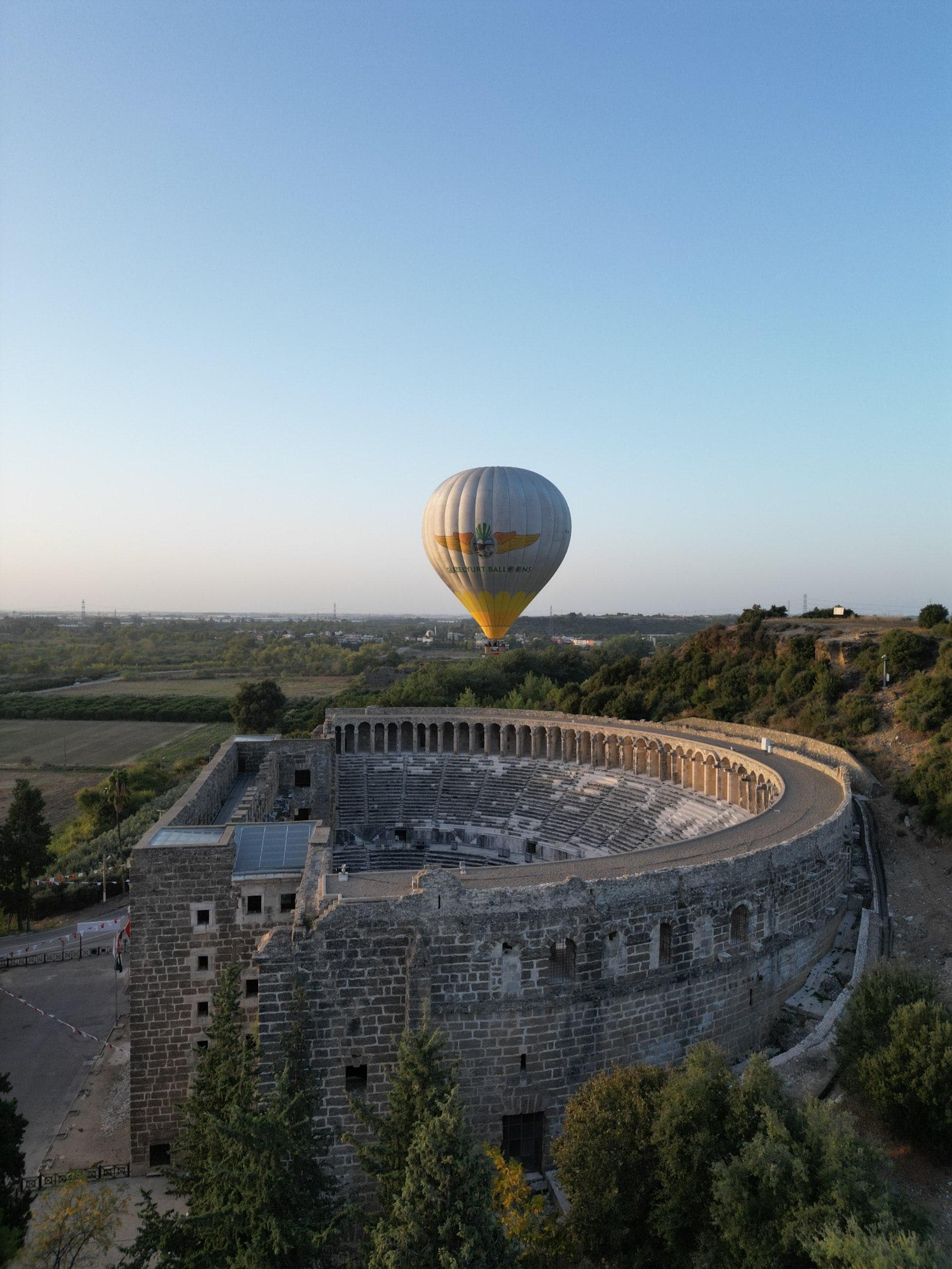 Antik Aspendos Tiyatrosu'nun taş sıralarının üzerinde gün doğumunda sarı-gri bir sıcak hava balonu süzülüyor
