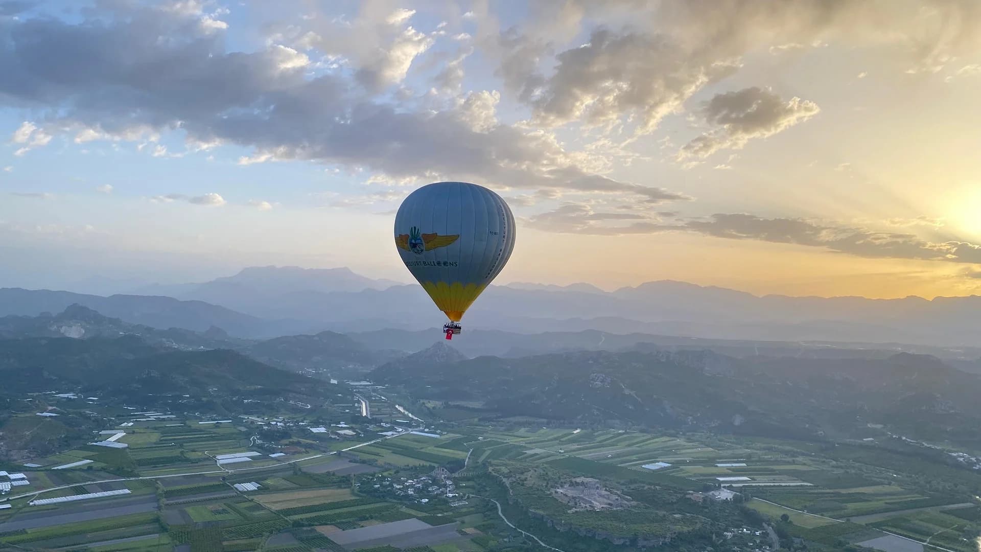Gün doğumunda dağların üzerinde alçalan bir sıcak hava balonu, aşağıda ova ve tarım arazileri uzanıyor