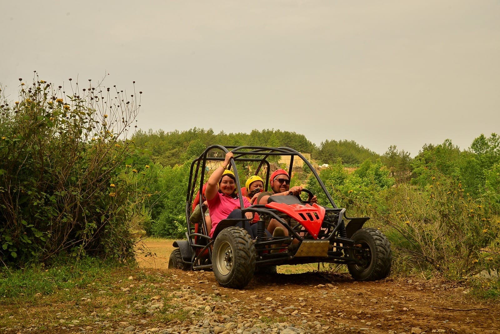 Kırmızı bir buggy'ye binen üç kişi kasklı gülümseyerek el sallıyor, arka planda tepedeki köy evleri