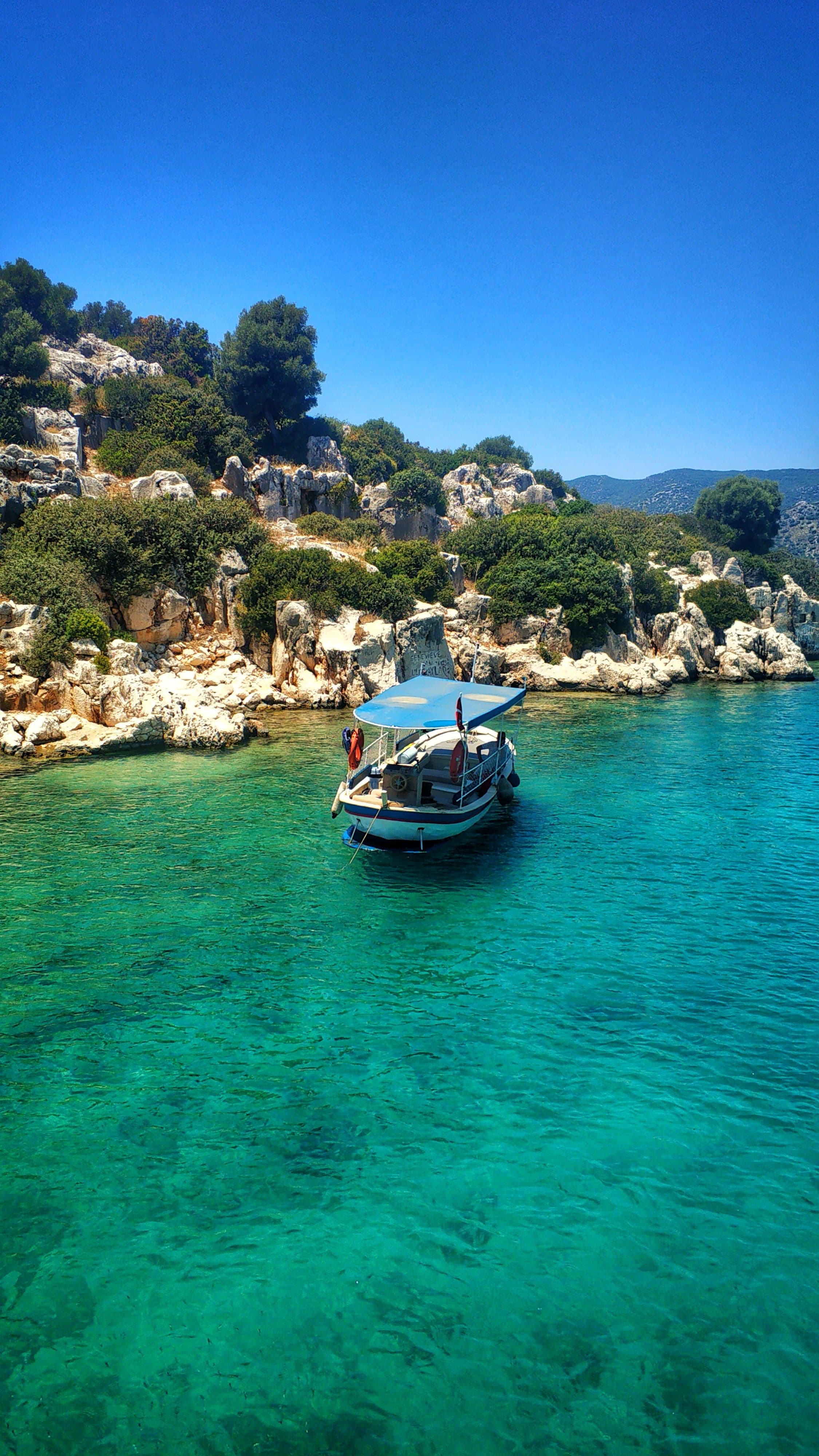 Mavi tenteli küçük bir tekne Kekova kıyısındaki kayalıkların önünde berrak turkuaz suda demirlemiş