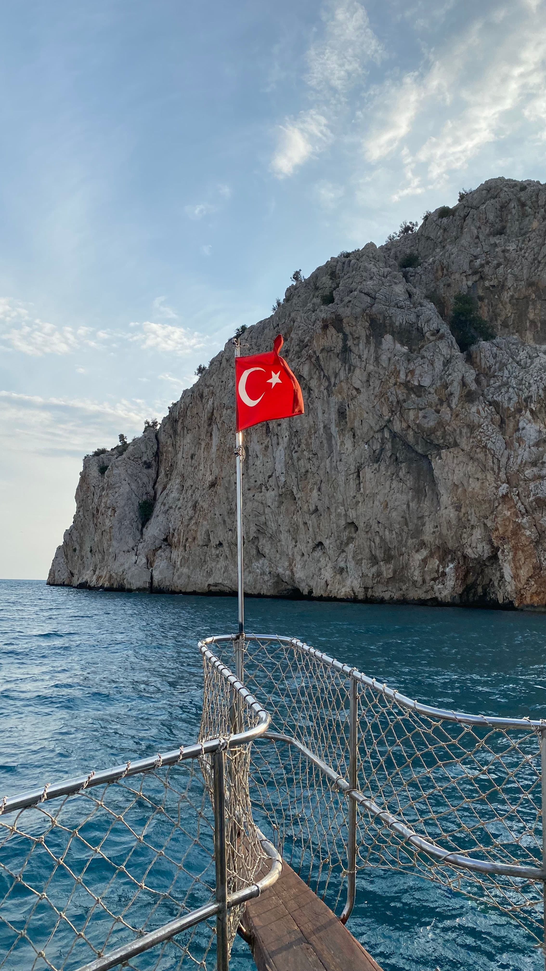 Teknenin pruvasında dalgalanan Türk bayrağı, arkada dik kayalık bir burun ve deniz