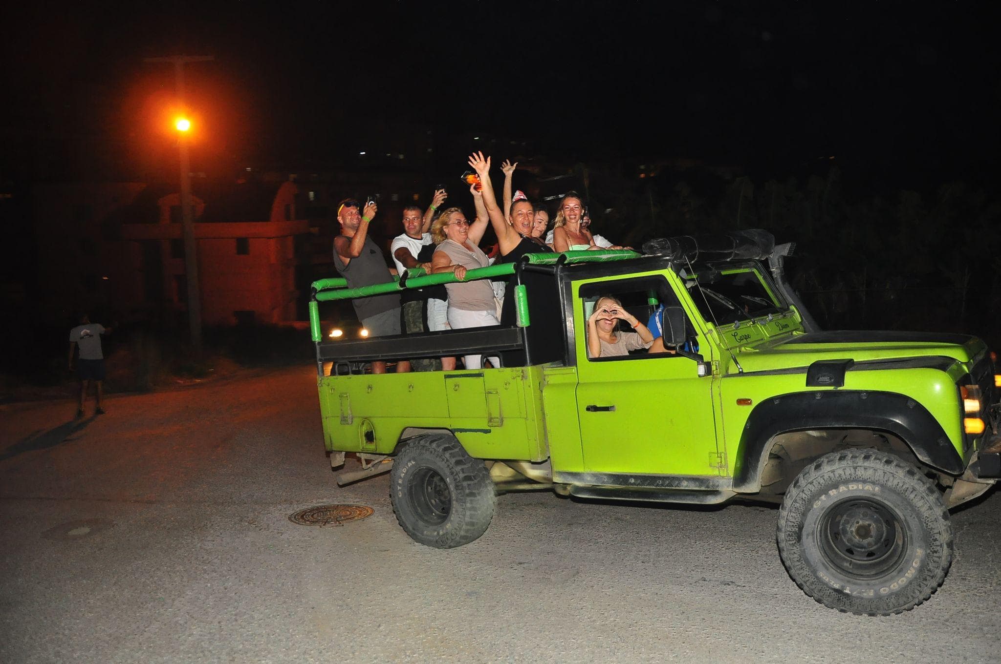 Yeşil üstü açık safari aracı gece sokakta ilerliyor, misafirler arkada ayakta kadeh kaldırıp el sallıyor