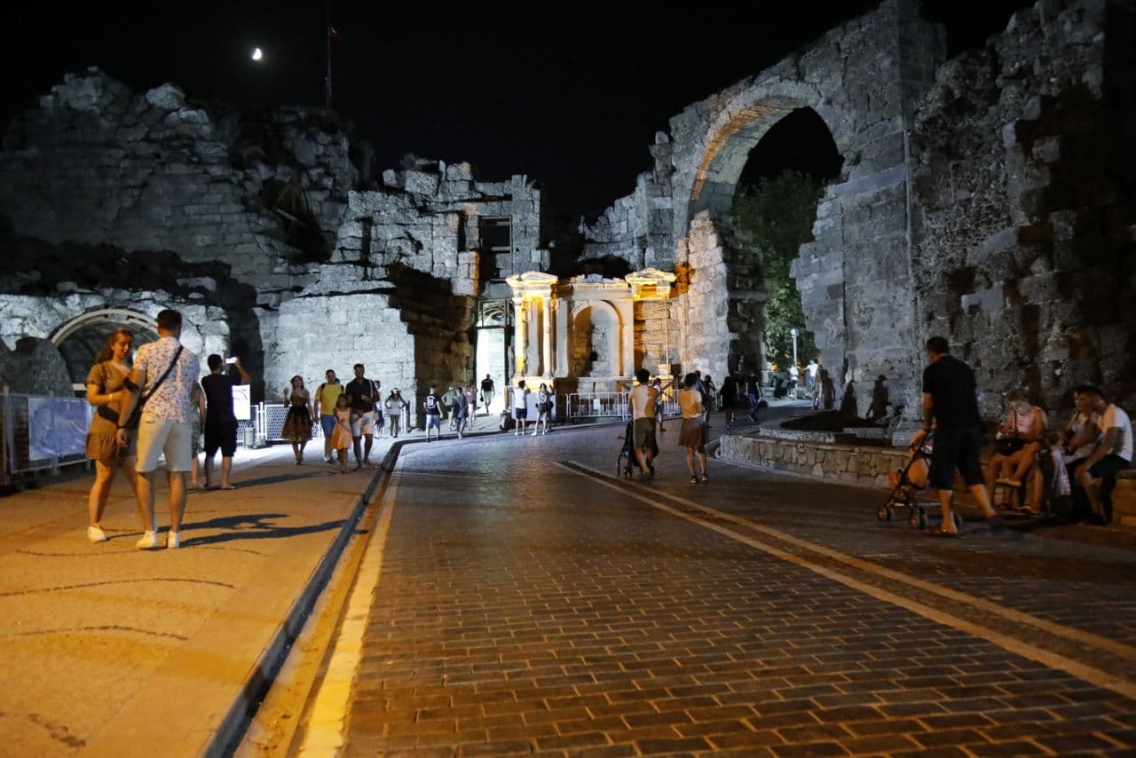 Gece Side'nin taş kemerli antik kapısı aydınlatılmış, önündeki taş döşeli yolda gezen ziyaretçiler