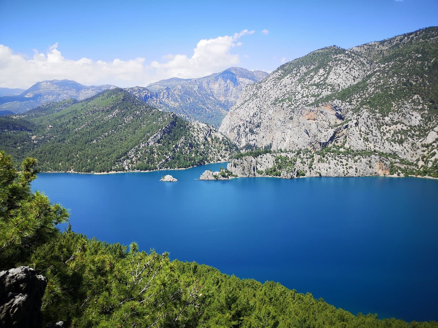 Yeşil ormanlarla çevrili derin mavi bir baraj gölü dağların arasında kıvrılıyor, ortasında küçük kayalık adacıklar var