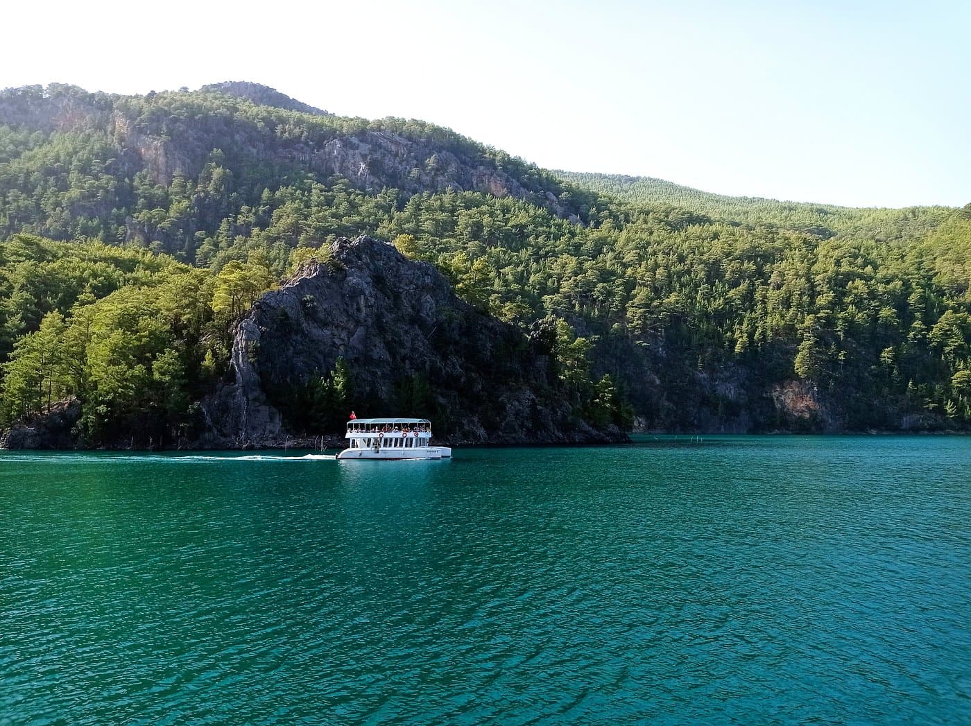 Tur teknesi ormanlık bir kayalık burnun önünden geçiyor, arkasında çam ağaçlarıyla kaplı dağlar ve turkuaz göl suyu