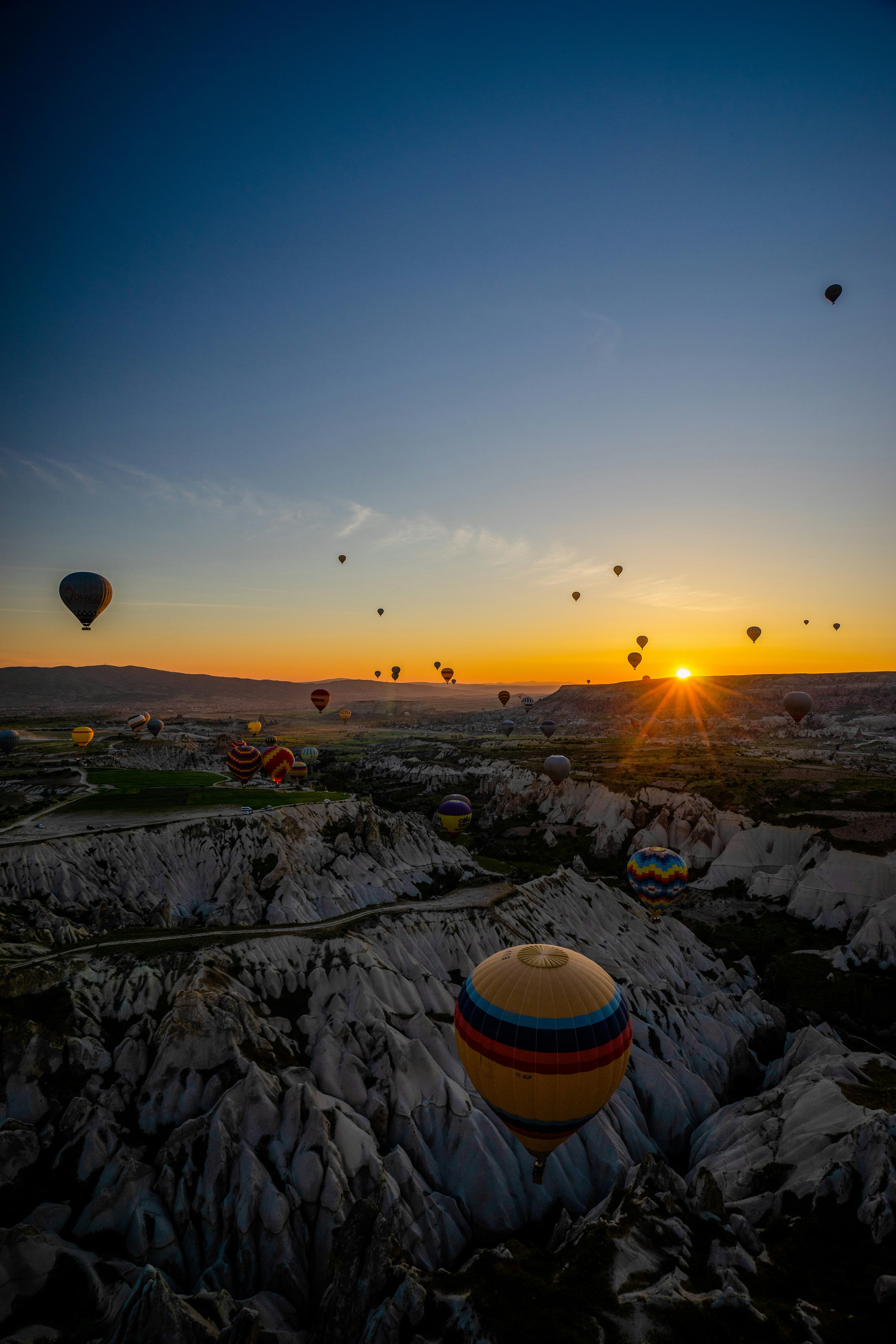 Gün doğarken sarı-kırmızı çizgili bir sıcak hava balonu kayalık vadinin hemen üzerinde alçaktan süzülüyor, arka planda ufukta güneş ve daha birçok balon