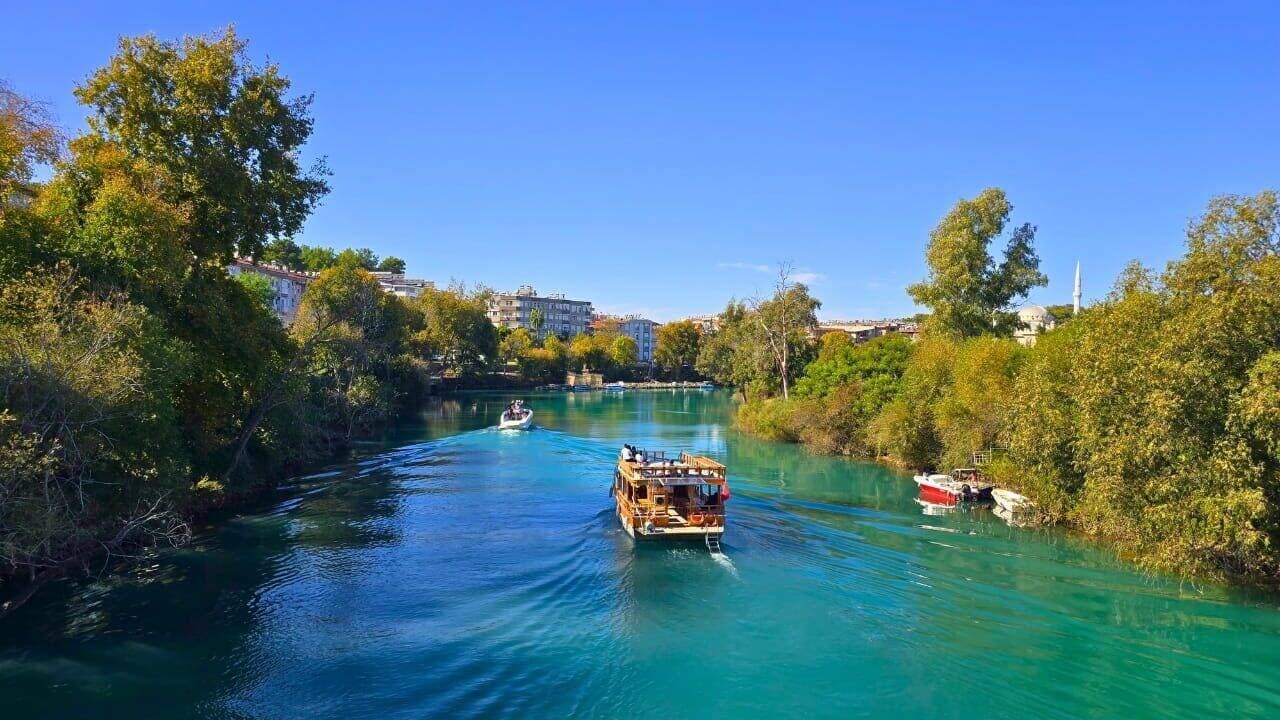 Turkuaz Manavgat Nehri'nde ahşap bir tur teknesi ilerliyor, iki kıyıda yeşil ağaçlar ve arka planda şehir binaları ile bir minare görünüyor