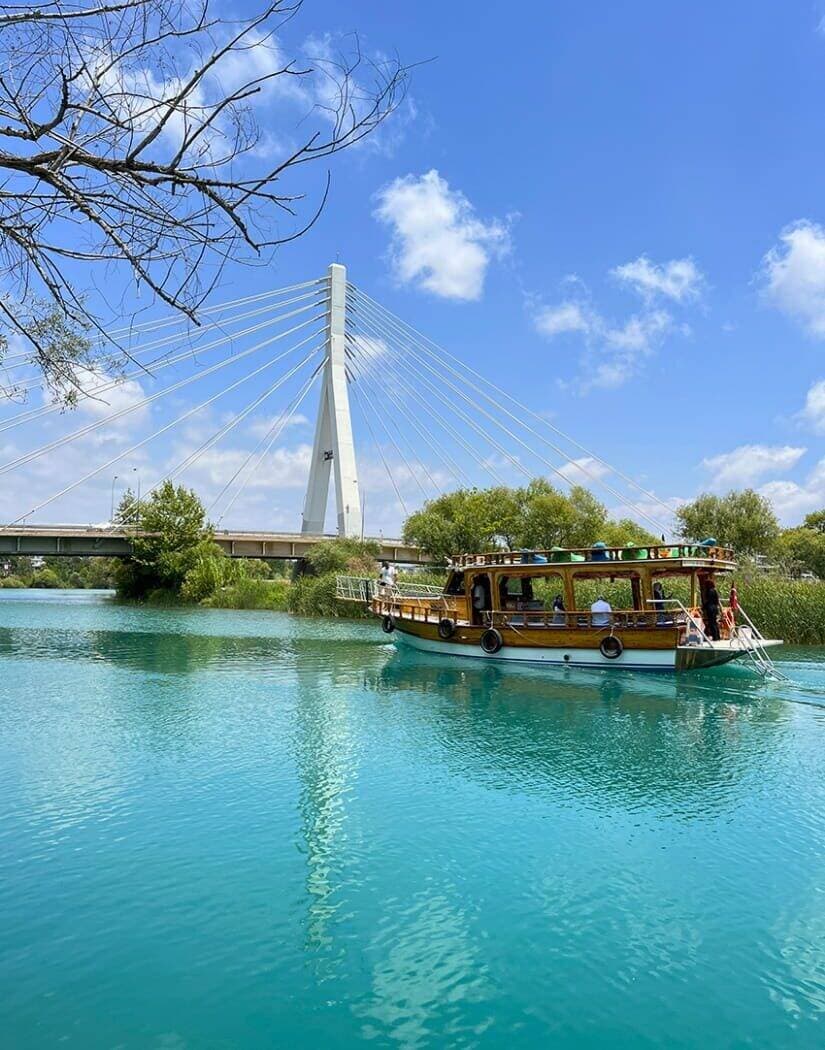 Ahşap tur teknesi Manavgat Nehri'nde asma köprünün önünden geçiyor, güvertede yolcular ayakta duruyor