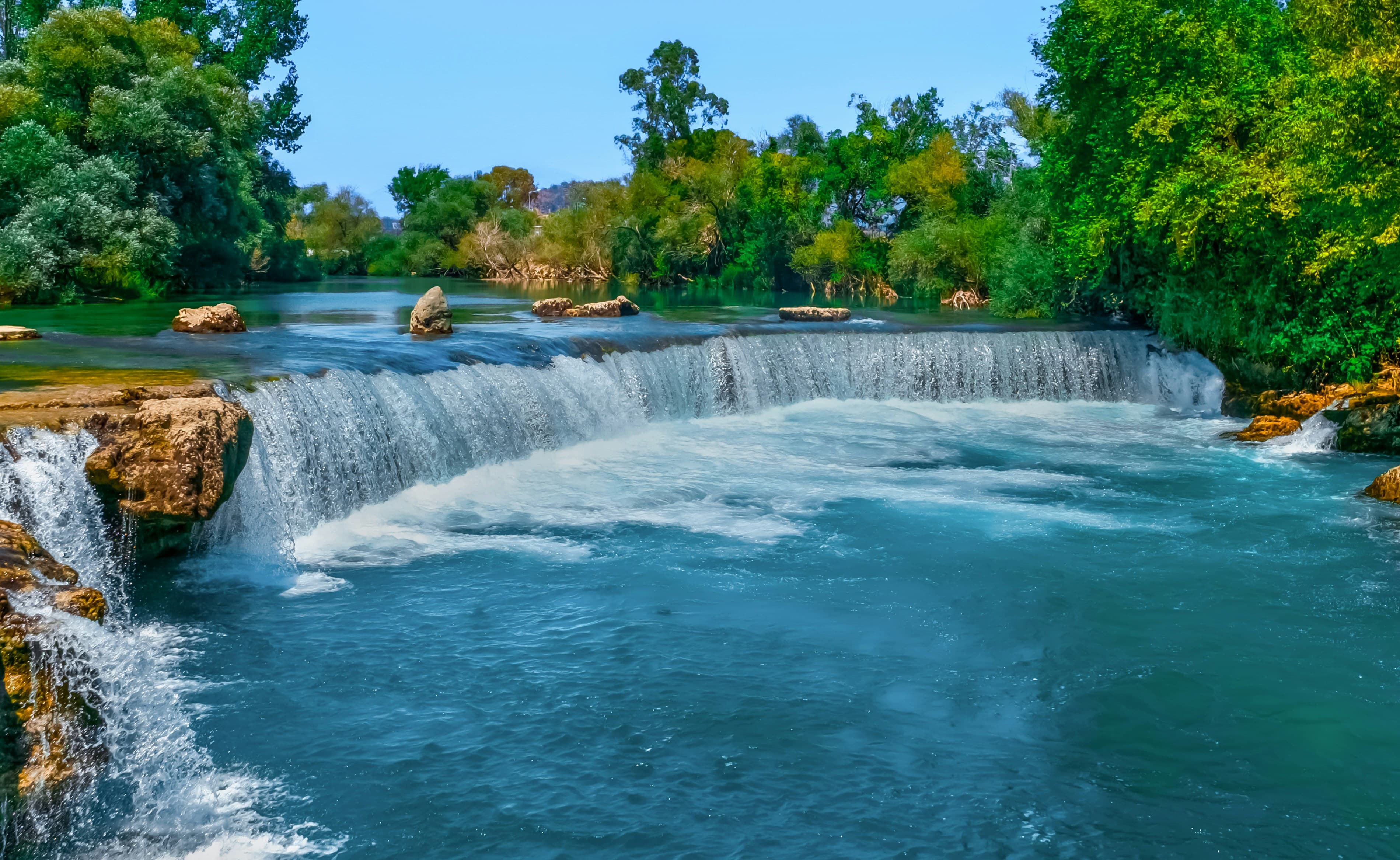 Manavgat Şelalesi'nin geniş beyaz köpüklü suyu kayalıklardan turkuaz nehre dökülüyor, çevresinde yemyeşil ağaçlar
