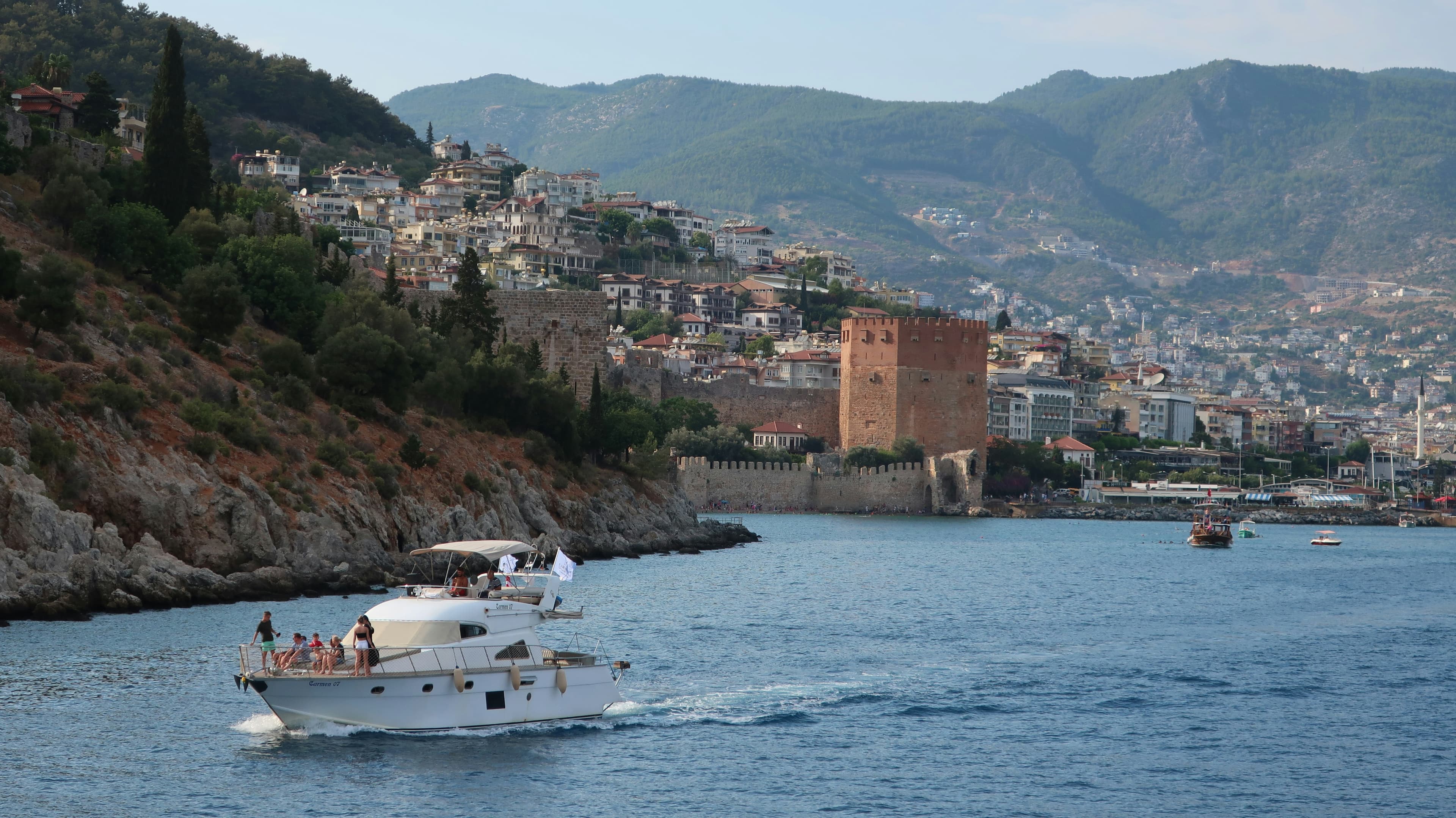 Kızıl Kule ve Alanya Kalesi surlarının önünden geçen beyaz bir motoryat, güvertesinde birkaç yolcu var