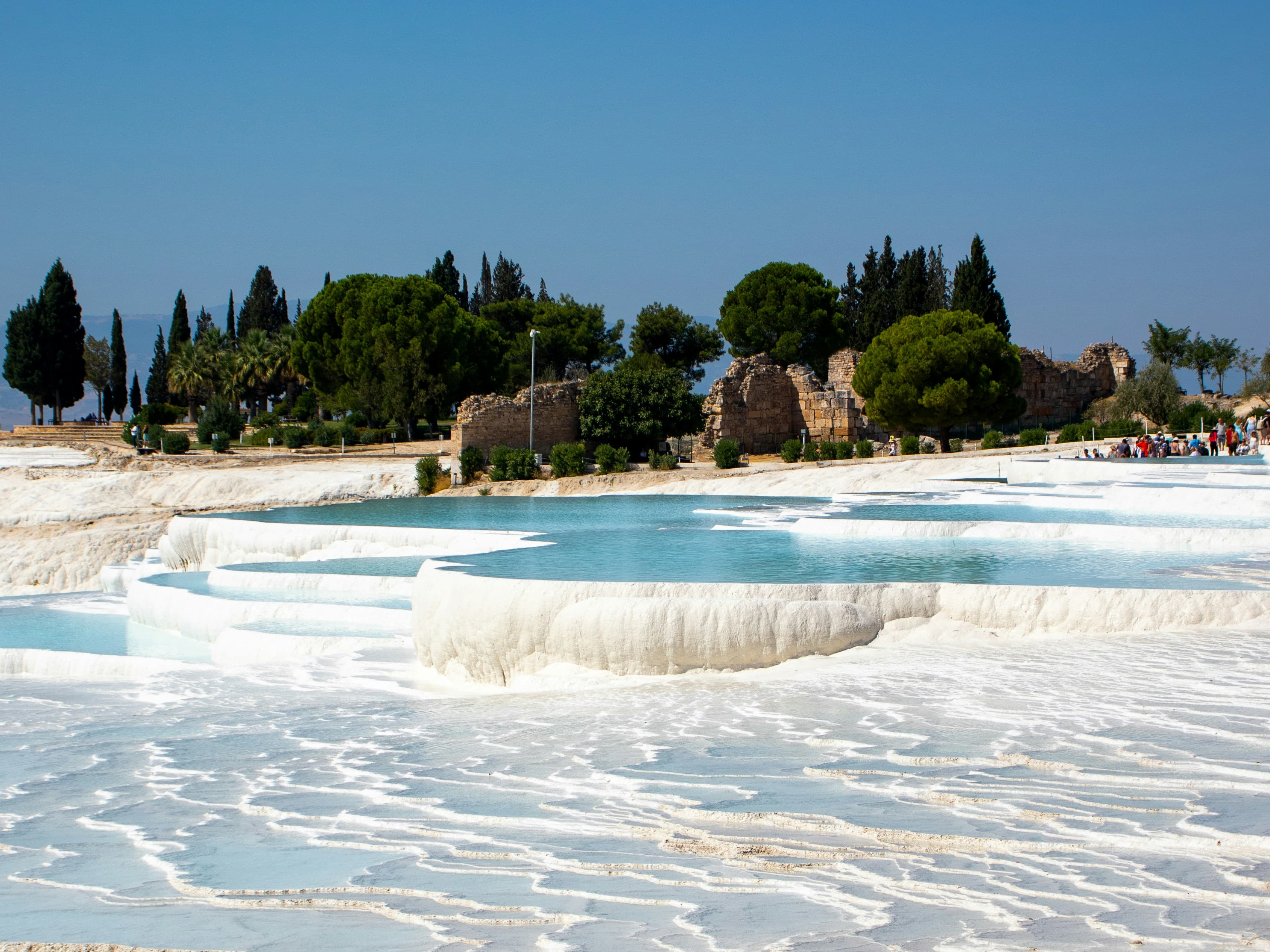 Pamukkale'nin beyaz travertenlerindeki turkuaz sıcak su havuzları, arkada antik kalıntılar ve servi ağaçları