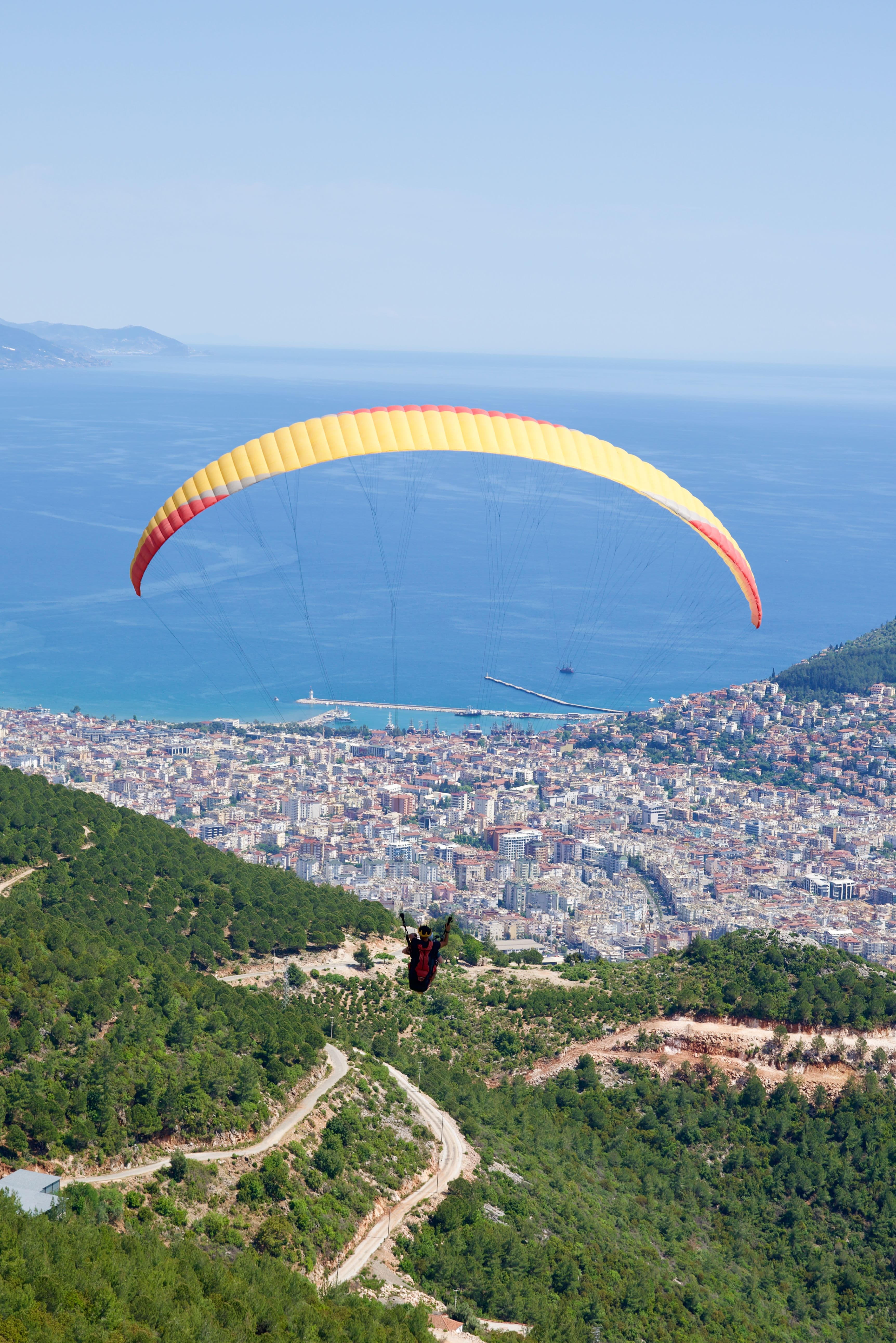 Tandem yamaç paraşütçüsü kalkıştan hemen sonra Alanya şehrinin ve limanının üzerinde süzülüyor, sarı-kırmızı şemsiye arkada denizle birlikte görünüyor