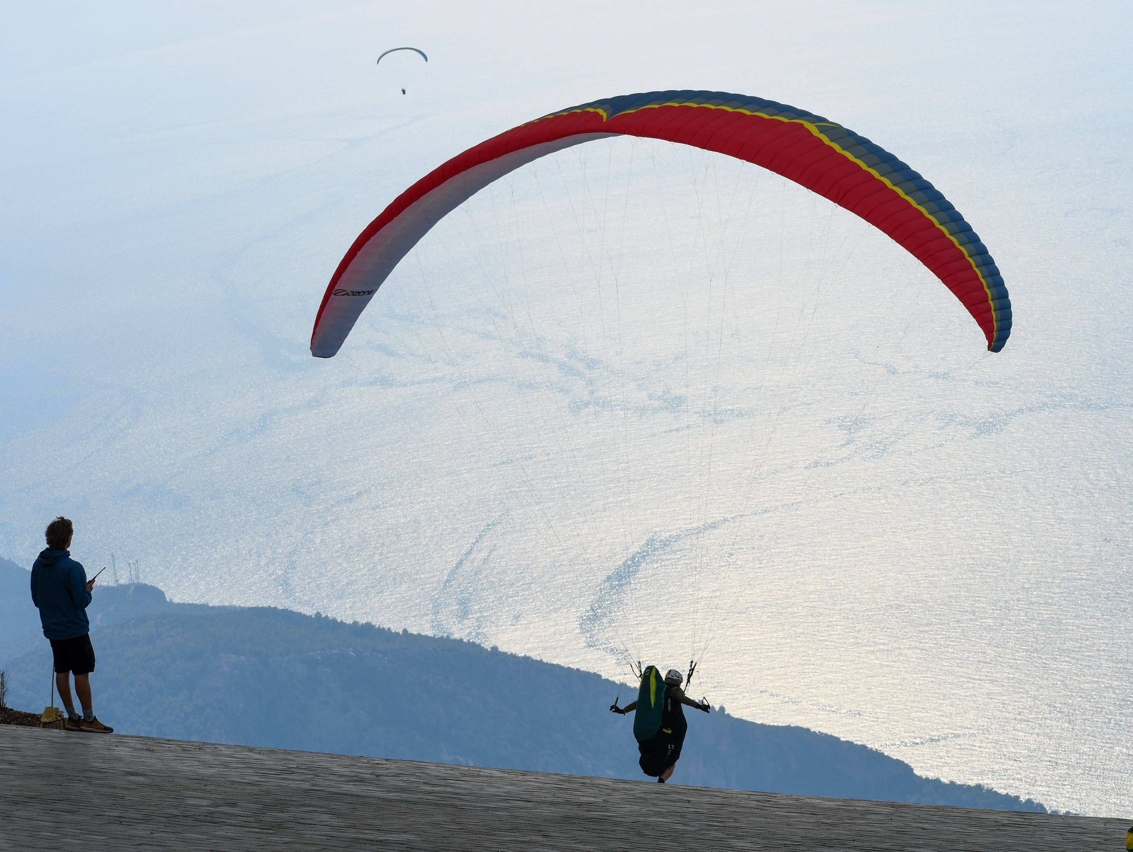 Yassı Tepe'deki kalkış pistinde kırmızı-mavi bir yamaç paraşütü açılmış, pilot koşarak havalanıyor, arkada uçan başka bir paraşütçü ve deniz manzarası