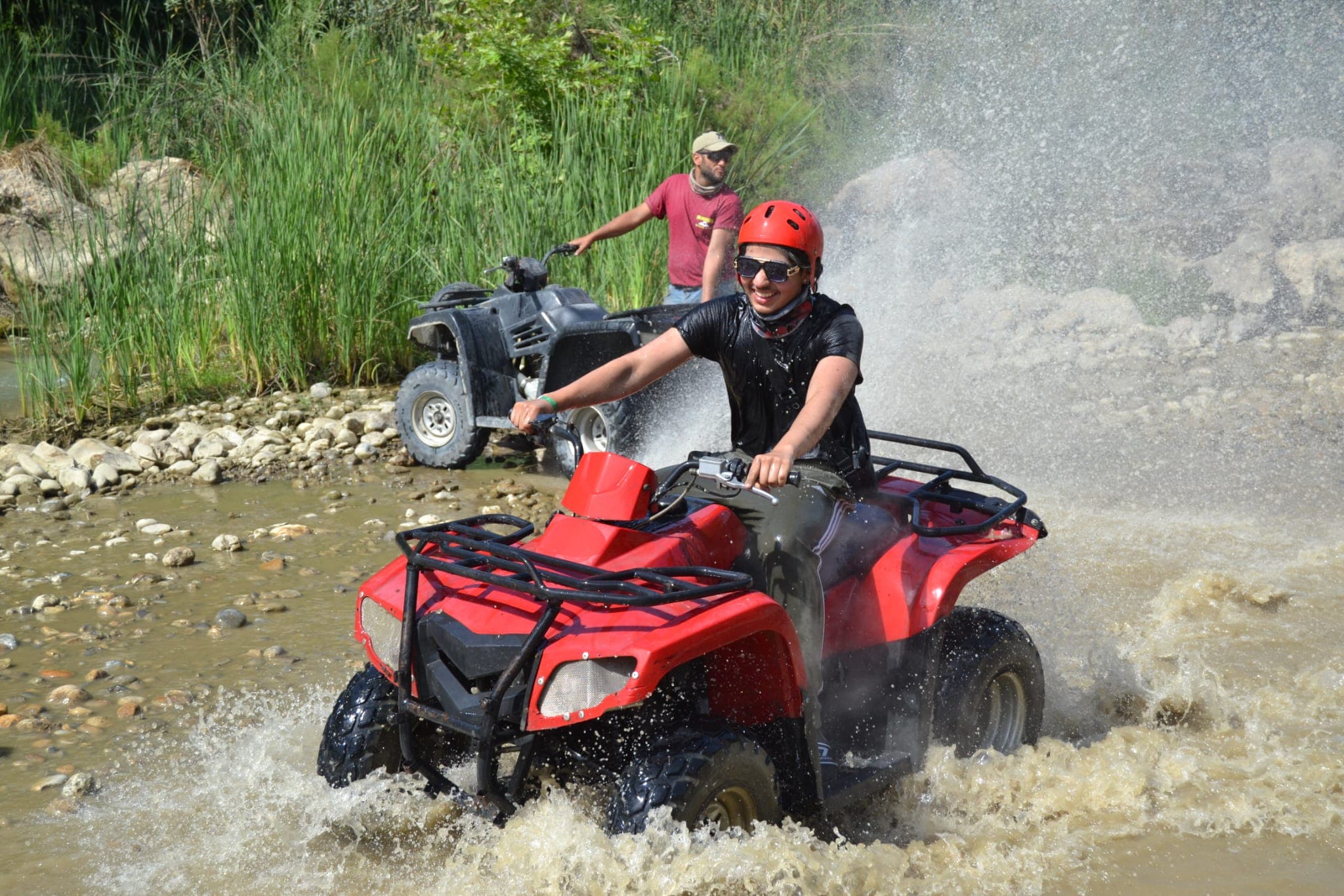 Kırmızı kasklı bir sürücü ATV'siyle dereden geçerken su sıçratıyor, kenarda rehber bekliyor