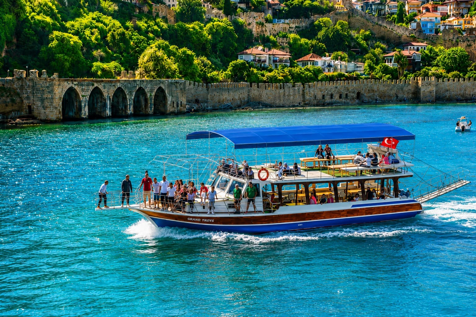 Mavi tenteli tekne, Alanya'nın kemerli su kemeri ve kale surlarının önünden geçiyor; pruvada ayakta yolcular, kıçta Türk bayrağı dalgalanıyor