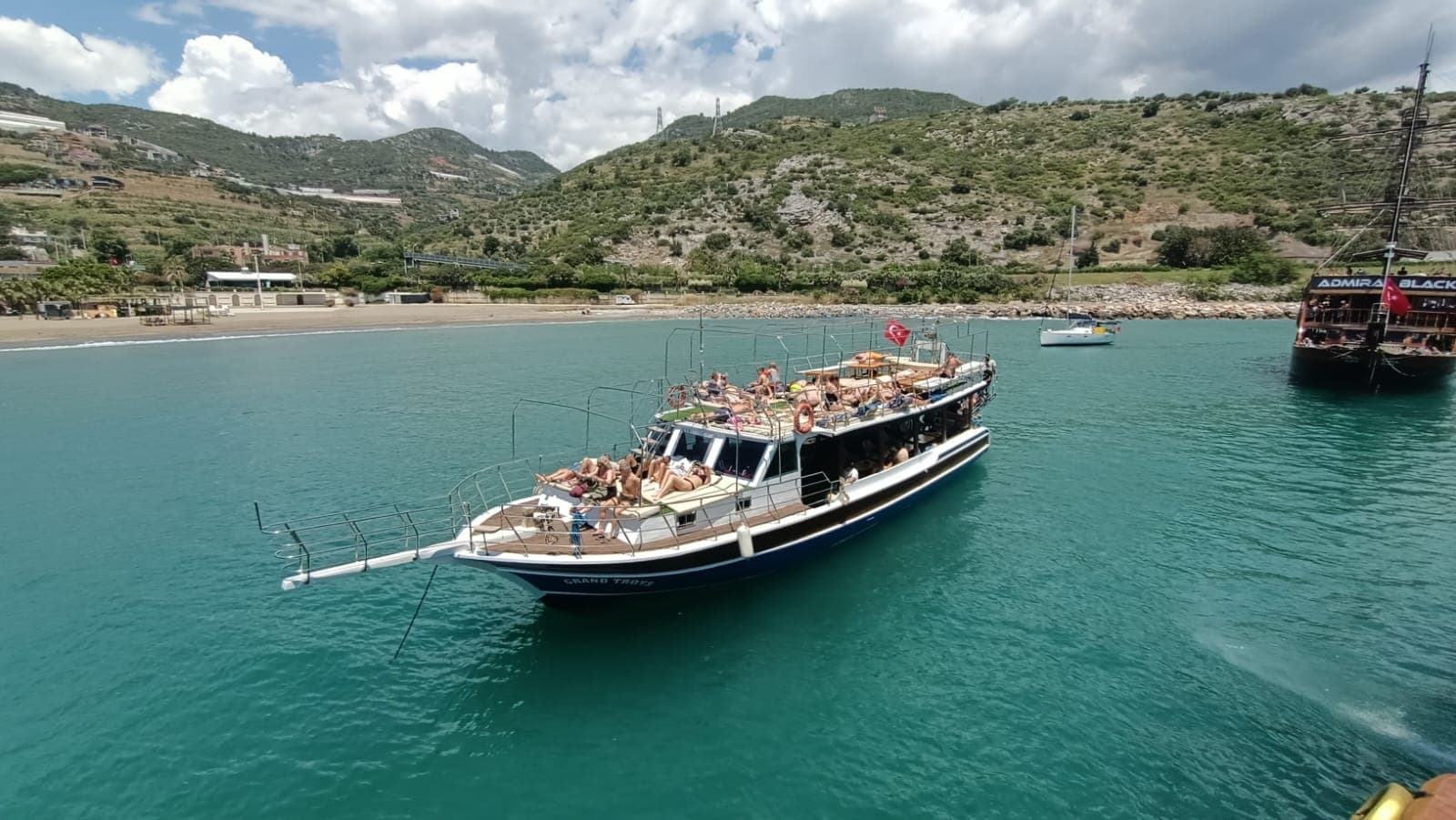 Bej tenteli tekne bir koyda demirlemiş; pruvada güneşlenen yolcular, arka planda dağlar, yakınında bir yelkenli ve siyah gövdeli büyük bir tekne, gökyüzünde bulutlar