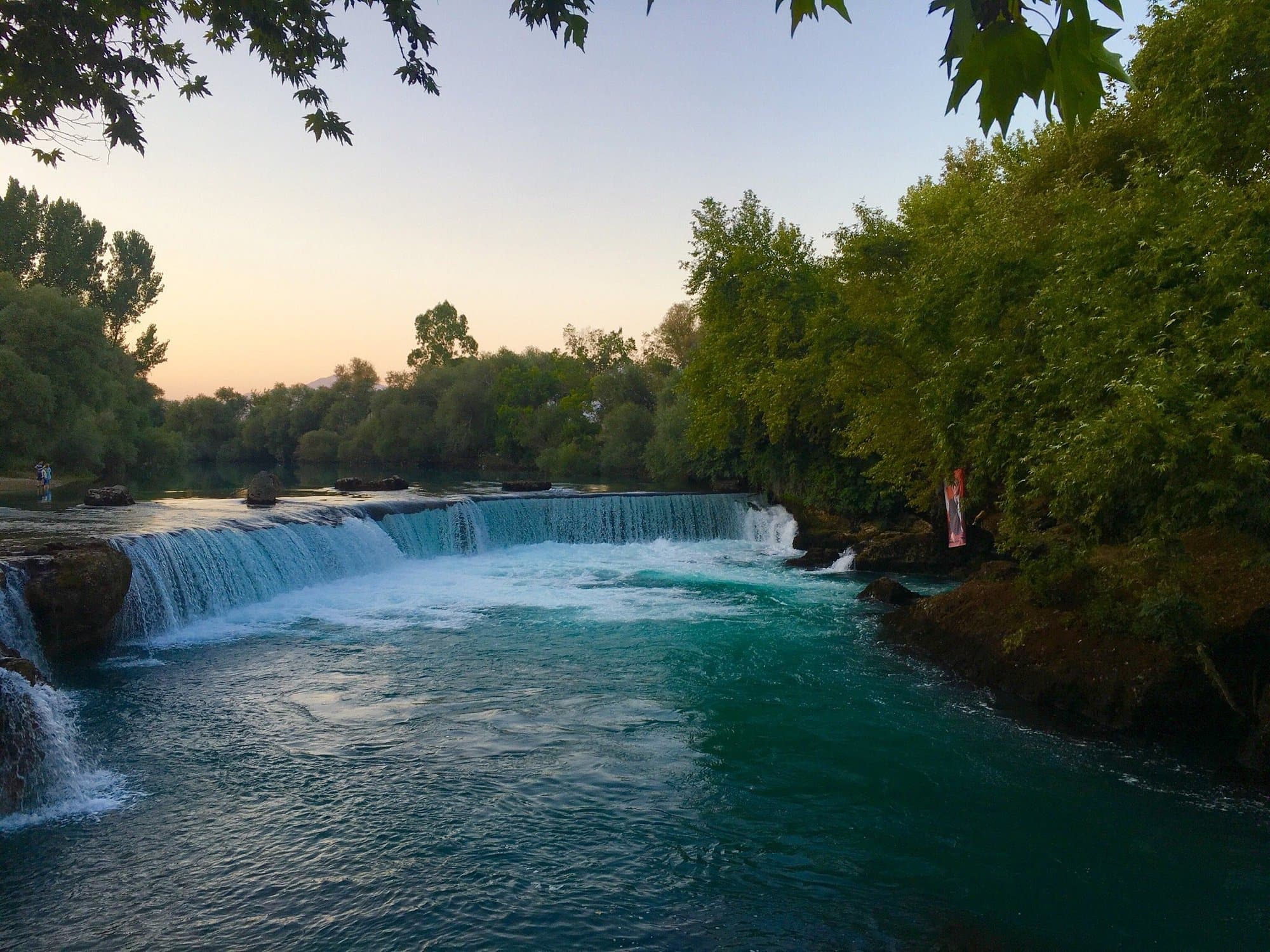 Manavgat Şelalesi'nin geniş beyaz köpüklü suyu kayalıklardan turkuaz nehre dökülüyor, iki yanı yeşil ağaçlarla çevrili