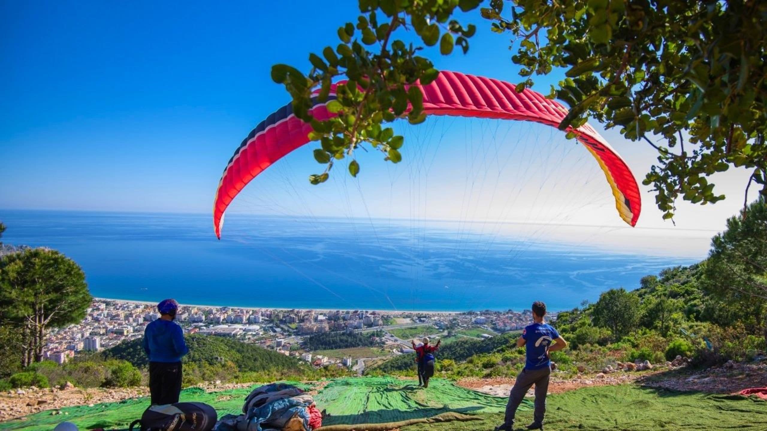 Kalkış pistinde açılmış kırmızı bir yamaç paraşütünün altında iki eğitmen dururken, uzakta bir başka paraşütçü Alanya kıyısı ve şehrinin üzerinde süzülüyor