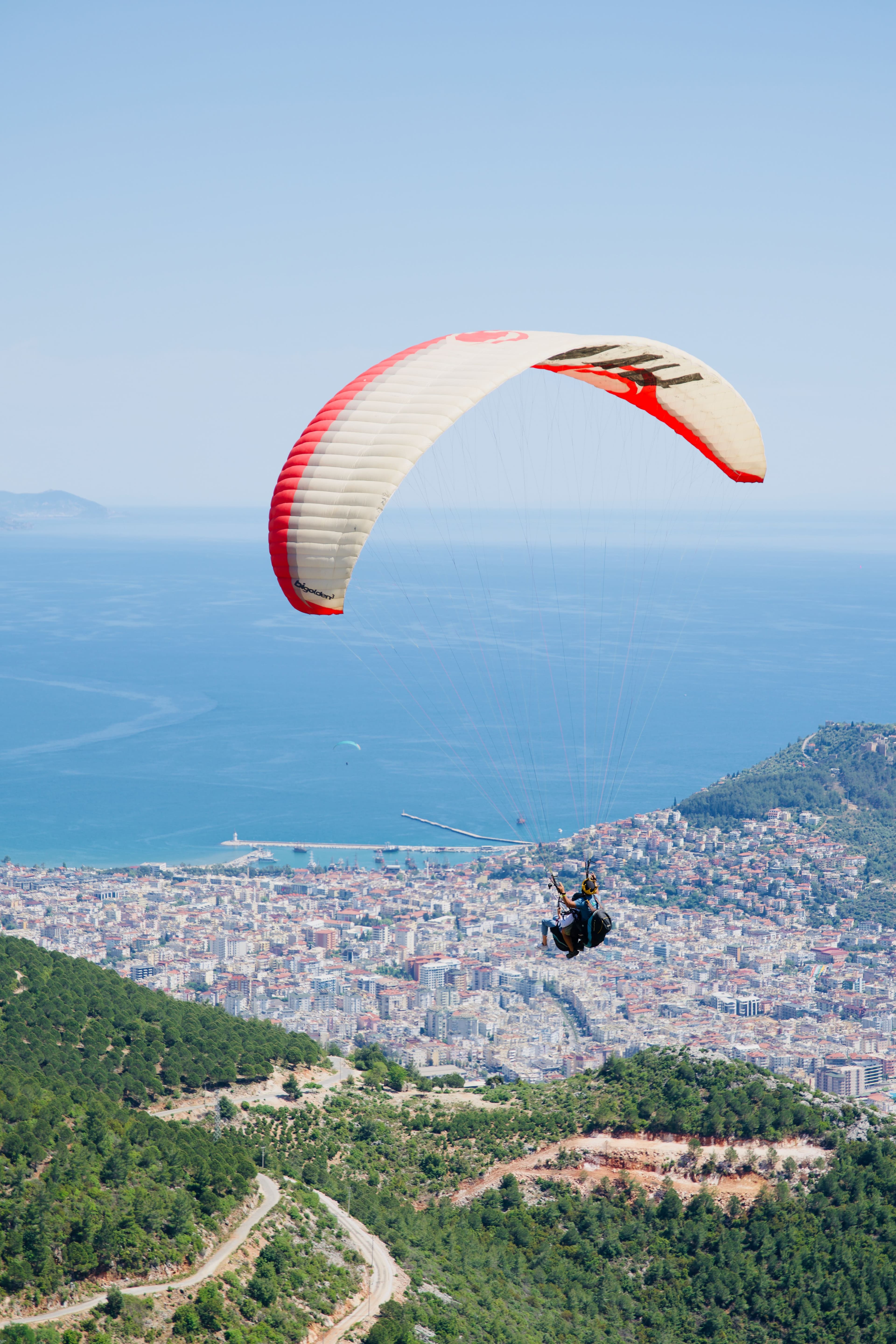 Tandem yamaç paraşütü Alanya limanı ve şehrinin üzerinde alçalırken kırmızı-beyaz şemsiye net görünüyor, arkada uzakta bir kara parçası