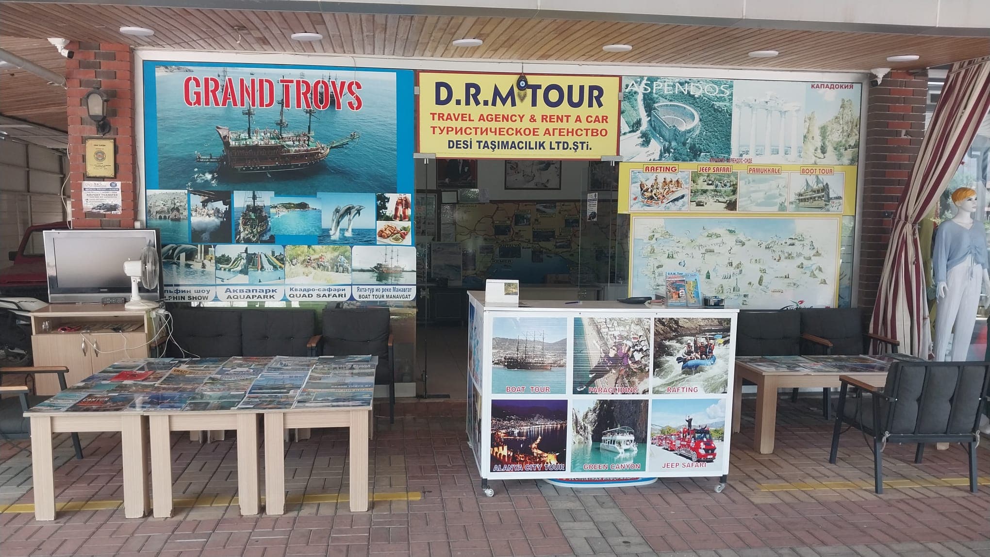 D.R.M. Tour ofisinin sokaktan görünümü: camlı cephede sarı 'D.R.M. TOUR' tabelası, duvarlarda tur afişleri, önünde broşür masaları ve oturma grubu